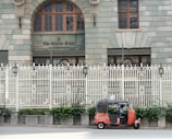 A red and black auto rickshaw is parked on the side of a street in front of a large, ornate building with arched windows and a decorative white fence. The building features a sign reading 'The Central Point' above the entrance. Greenery grows at the base of the fence, adding a touch of nature to the urban setting.