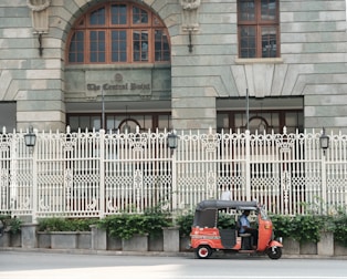 A red and black auto rickshaw is parked on the side of a street in front of a large, ornate building with arched windows and a decorative white fence. The building features a sign reading 'The Central Point' above the entrance. Greenery grows at the base of the fence, adding a touch of nature to the urban setting.