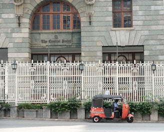 A red and black auto rickshaw is parked on the side of a street in front of a large, ornate building with arched windows and a decorative white fence. The building features a sign reading 'The Central Point' above the entrance. Greenery grows at the base of the fence, adding a touch of nature to the urban setting.