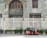 A red and black auto rickshaw is parked on the side of a street in front of a large, ornate building with arched windows and a decorative white fence. The building features a sign reading 'The Central Point' above the entrance. Greenery grows at the base of the fence, adding a touch of nature to the urban setting.