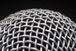 A close-up view of a metallic mesh structure with a grid-like pattern. The material appears to be interwoven, creating a textured and intricate design. The background is dark, highlighting the metallic surface.
