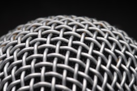A close-up view of a metallic mesh structure with a grid-like pattern. The material appears to be interwoven, creating a textured and intricate design. The background is dark, highlighting the metallic surface.