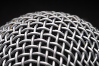 A close-up view of a metallic mesh structure with a grid-like pattern. The material appears to be interwoven, creating a textured and intricate design. The background is dark, highlighting the metallic surface.