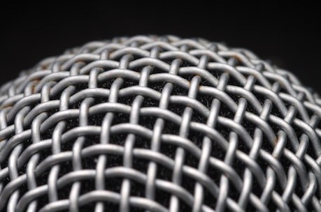 A close-up view of a metallic mesh structure with a grid-like pattern. The material appears to be interwoven, creating a textured and intricate design. The background is dark, highlighting the metallic surface.