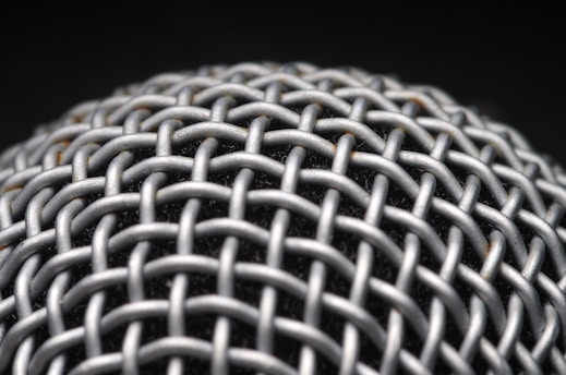 A close-up view of a metallic mesh structure with a grid-like pattern. The material appears to be interwoven, creating a textured and intricate design. The background is dark, highlighting the metallic surface.