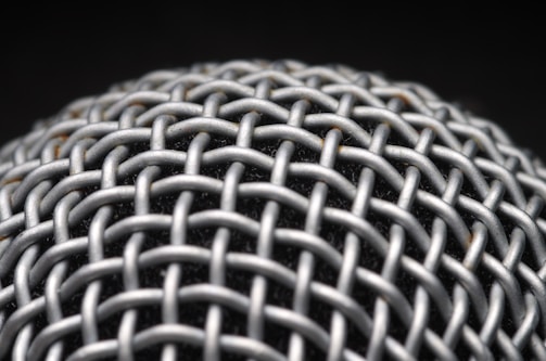A close-up view of a metallic mesh structure with a grid-like pattern. The material appears to be interwoven, creating a textured and intricate design. The background is dark, highlighting the metallic surface.