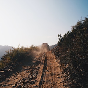 A rugged off-road vehicle travels along a dusty, unpaved path bordered by wild vegetation. The scene conveys a sense of adventure and remoteness under a clear blue sky.
