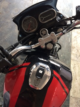 A close-up view of a motorcycle's dashboard and fuel tank, featuring a speedometer, tachometer, and handlebar controls. The fuel tank is red with black racing stripes and a shiny chrome cap.