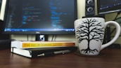 A workspace featuring a computer monitor displaying lines of code in blue and green on a dark background occupies the center. In the foreground, there is a white coffee mug with a black tree design placed on a wooden desk. Next to the mug, a closed notebook and a book titled 'The Art of Learning' by Josh Waitzkin are laid out. A pen rests on top of the notebook.