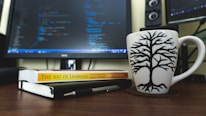 A workspace featuring a computer monitor displaying lines of code in blue and green on a dark background occupies the center. In the foreground, there is a white coffee mug with a black tree design placed on a wooden desk. Next to the mug, a closed notebook and a book titled 'The Art of Learning' by Josh Waitzkin are laid out. A pen rests on top of the notebook.