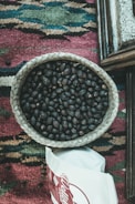 A woven basket is filled with dark roasted coffee beans, placed on a multi-colored woven textile with geometric patterns. Nearby, there is a partially visible white bag with red lettering.