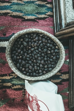 A woven basket is filled with dark roasted coffee beans, placed on a multi-colored woven textile with geometric patterns. Nearby, there is a partially visible white bag with red lettering.