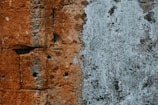 A textured surface displaying a wall with a blend of materials. The left side features reddish-brown stone blocks with visible drill holes, showing weathering and age. The right side reveals a patchy surface coated with a whitish, peeling layer suggesting layers of paint or plaster.