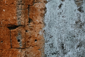 A textured surface displaying a wall with a blend of materials. The left side features reddish-brown stone blocks with visible drill holes, showing weathering and age. The right side reveals a patchy surface coated with a whitish, peeling layer suggesting layers of paint or plaster.