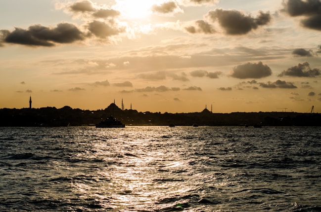 A sunset over a large body of water with silhouettes of buildings and minarets in the background. The sky is partly cloudy, casting reflections on the rippled water below.