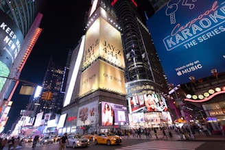 A bustling city intersection at night is illuminated by bright billboards and advertisements. Yellow taxis and numerous pedestrians fill the streets, highlighting the urban vibrancy. Prominent advertisements include a music release and a show promotion, contributing to the lively atmosphere.
