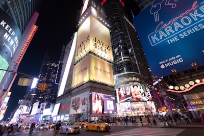A bustling city intersection at night is illuminated by bright billboards and advertisements. Yellow taxis and numerous pedestrians fill the streets, highlighting the urban vibrancy. Prominent advertisements include a music release and a show promotion, contributing to the lively atmosphere.