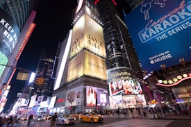 A bustling city intersection at night is illuminated by bright billboards and advertisements. Yellow taxis and numerous pedestrians fill the streets, highlighting the urban vibrancy. Prominent advertisements include a music release and a show promotion, contributing to the lively atmosphere.