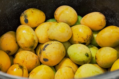 A collection of ripe mangoes with a mix of yellow and green colors, some showing dark spots, piled together in a black container.