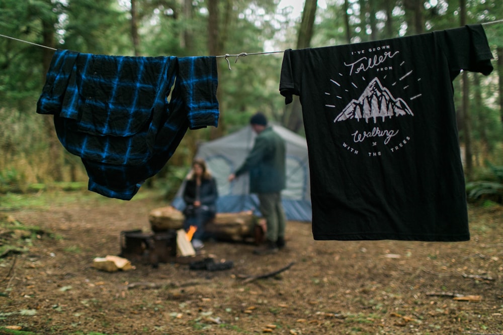 two shirts hanging near woman and man beside blue tent under tall trees