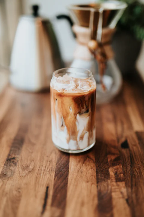 Tall iced coffee in a glass with foamy crema on a pale wooden cafe counter