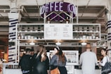 A busy coffee shop environment with a modern aesthetic. A group of people stand in front of a counter that displays a menu listing drinks and food options. The decor includes a neon 'Coffee' sign above the counter. Shelves are lined with coffee bags and various items, contributing to an industrial yet cozy atmosphere.