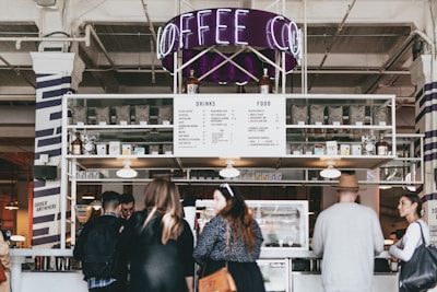 A busy coffee shop environment with a modern aesthetic. A group of people stand in front of a counter that displays a menu listing drinks and food options. The decor includes a neon 'Coffee' sign above the counter. Shelves are lined with coffee bags and various items, contributing to an industrial yet cozy atmosphere.