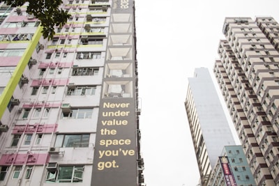 Tall buildings tower upwards with various patterns of windows and balconies. One building features a striking message in large, bold letters that reads 'Never under value the space you’ve got.' The scene includes a mix of architectural styles and details, with pink, green, and brown color accents. A tree's leaves are partially visible in the foreground, adding a touch of nature to the urban environment.