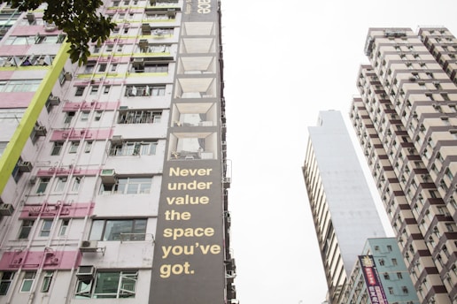 Tall buildings tower upwards with various patterns of windows and balconies. One building features a striking message in large, bold letters that reads 'Never under value the space you’ve got.' The scene includes a mix of architectural styles and details, with pink, green, and brown color accents. A tree's leaves are partially visible in the foreground, adding a touch of nature to the urban environment.