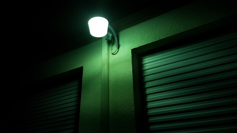 An outdoor wall-mounted light illuminates a building with a greenish glow. The wall features a textured surface, and two metal roll-up doors are visible, creating a crisp contrast with the shadows.