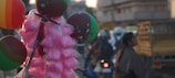 A collection of colorful balloons and bags of pink cotton candy are displayed prominently on a pole. In the background, there is a busy street scene with several blurred figures, vehicles, and buildings. The atmosphere appears lively and vibrant.