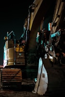 A large yellow excavator with visible tracks and an arm is positioned in a dimly lit outdoor setting, with its metal components reflecting a subtle glow from nearby lights.