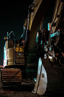A large yellow excavator with visible tracks and an arm is positioned in a dimly lit outdoor setting, with its metal components reflecting a subtle glow from nearby lights.