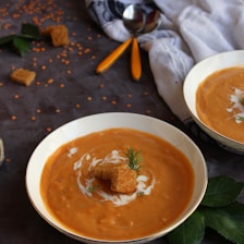 A bowl of creamy orange soup topped with croutons and a sprig of green herb, placed on a dark table cloth. Nearby, there are scattered lentils, two spoons with yellow handles, and another bowl of soup partially visible. A white and black patterned cloth adds a cozy touch to the setting.