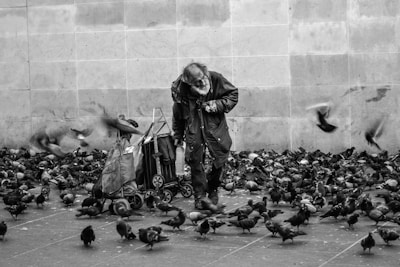 grayscale photography of man surrounded by flock of pigeons standing on street