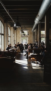 The spacious interior of a cafe with large windows allowing natural light to stream in, casting shadows across the floor. Customers are seated on sofas and chairs, engaging in conversations and enjoying their time. The decor features exposed ductwork, stone walls, and industrial-style lighting fixtures hanging from a high ceiling.