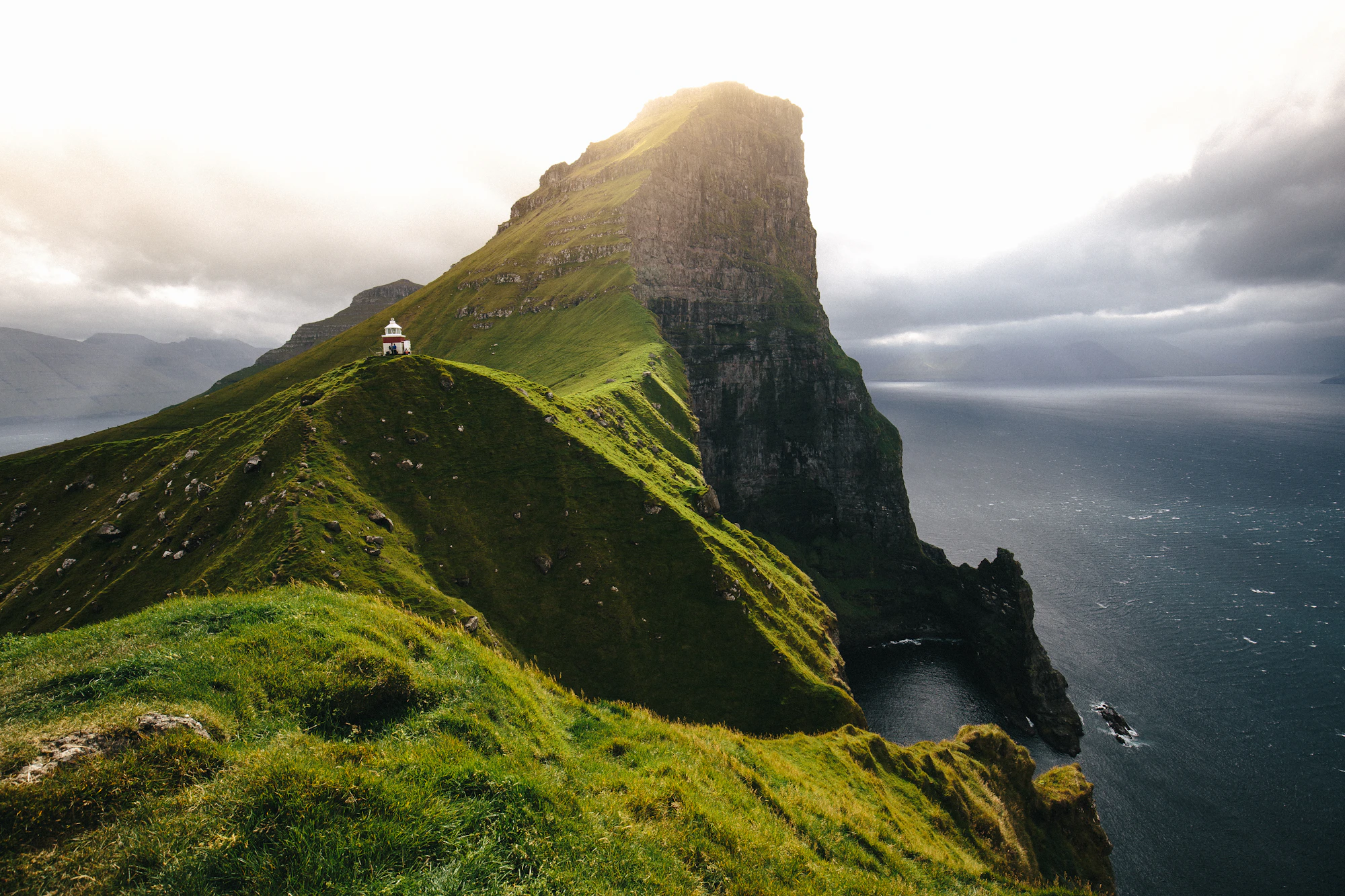 Kallur lighthouse on a green Kalsoy ridge, sunlight breaking over the dark sea