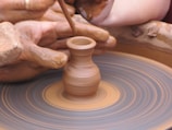 A close-up view of hands skillfully shaping a small clay pot on a spinning pottery wheel. The hands are covered in wet clay, and there is a sense of motion from the spinning wheel. The scene captures the intricate process of pottery making.