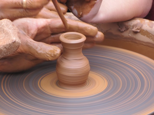 A close-up view of hands skillfully shaping a small clay pot on a spinning pottery wheel. The hands are covered in wet clay, and there is a sense of motion from the spinning wheel. The scene captures the intricate process of pottery making.