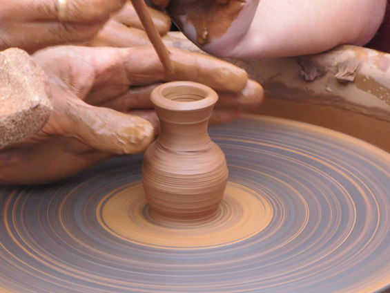 A close-up shot of a student's hands shaping clay on a spinning pottery wheel, with shelves of colorful glazes in the background.