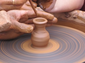 A close-up view of hands skillfully shaping a small clay pot on a spinning pottery wheel. The hands are covered in wet clay, and there is a sense of motion from the spinning wheel. The scene captures the intricate process of pottery making.