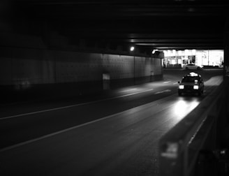 A taxi is driving through a dimly lit underpass with lights from the street and buildings visible in the distance. The road ahead is mostly empty, and the overall scene is captured in black and white.