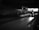 A taxi is driving through a dimly lit underpass with lights from the street and buildings visible in the distance. The road ahead is mostly empty, and the overall scene is captured in black and white.