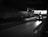 A taxi is driving through a dimly lit underpass with lights from the street and buildings visible in the distance. The road ahead is mostly empty, and the overall scene is captured in black and white.