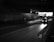 A taxi is driving through a dimly lit underpass with lights from the street and buildings visible in the distance. The road ahead is mostly empty, and the overall scene is captured in black and white.