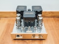 A vintage-style audio amplifier with vacuum tubes sits on a wooden floor. The device features several reflective metallic surfaces and cylindrical components housed in a rectangular chassis. A power switch and various knobs are present on the front panel.