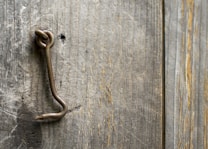 A rustic, textured wooden surface with vertical grain patterns and natural weathering. An antique metal hook is embedded into the wood, showing signs of rust and age. The hook is bent, suggesting past use or wear.