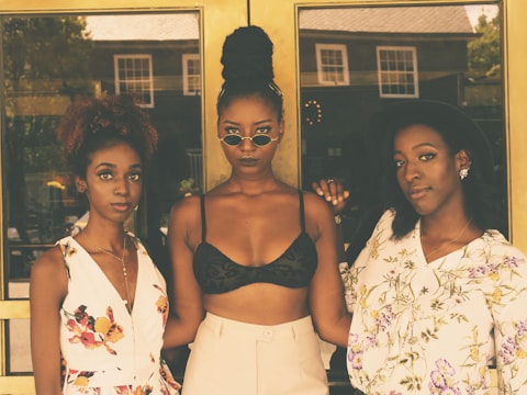 Three women are standing in front of a building with glass doors. The woman on the left is wearing a white floral dress, the woman in the middle is wearing a black crop top and light-colored pants, and the woman on the right is in a floral blouse and a black hat. They all pose closely together, confidently looking at the camera.