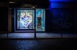 A tattoo parlor with a brightly lit neon sign in the window advertising 'Cesar Tattoos Lopez' in a heart shape with a crown on top. The shop interior is visible through the window, showing a chair and a colorful piece of artwork on a stand. The exterior of the shop is dark, with blue lighting creating a contrasting atmosphere.