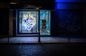 A tattoo parlor with a brightly lit neon sign in the window advertising 'Cesar Tattoos Lopez' in a heart shape with a crown on top. The shop interior is visible through the window, showing a chair and a colorful piece of artwork on a stand. The exterior of the shop is dark, with blue lighting creating a contrasting atmosphere.
